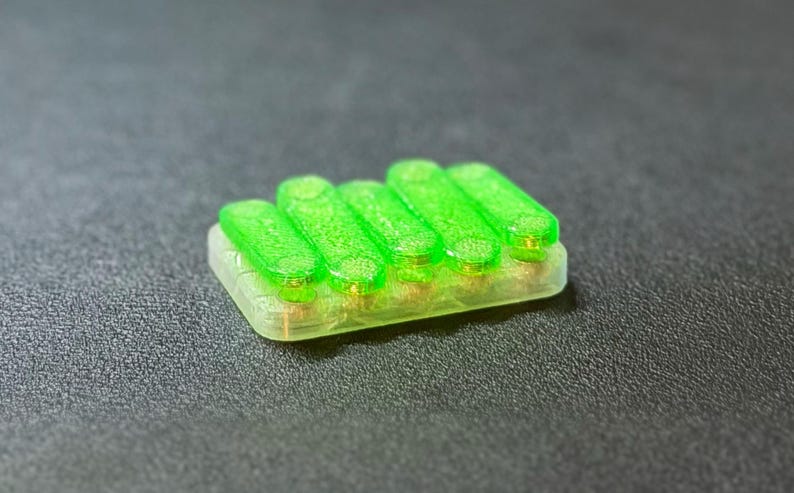 May include: A small, rectangular object with five bright green, cylindrical components arranged in a row. The base is a translucent, light green color. The object is set against a dark gray background.