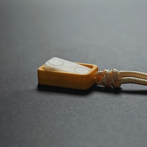 Puede incluir: Primer plano de un pequeño recipiente rectangular amarillo con un inserto blanco estampado. Una cuerda de color crema está unida al recipiente. El fondo es una superficie gris oscuro.