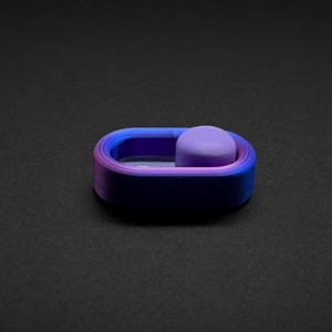 May include: An oval-shaped fidget slider featuring a gradient color scheme that transitions from deep purple to vibrant blue. The device includes a smooth, rounded purple button positioned on a track, allowing for back-and-forth sliding motion.