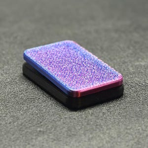 Puede incluir: Una caja rectangular negra con una parte superior brillante y con purpurina. La superficie superior tiene una transición de color de azul a morado a rosa. El artículo está sobre un fondo gris texturizado.