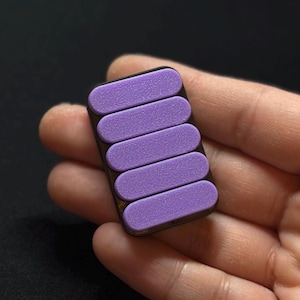 May include: A rectangular, dark brown object with five parallel, light purple, textured, rectangular pads. The object is held in a person's hand against a dark background. The pads are evenly spaced.