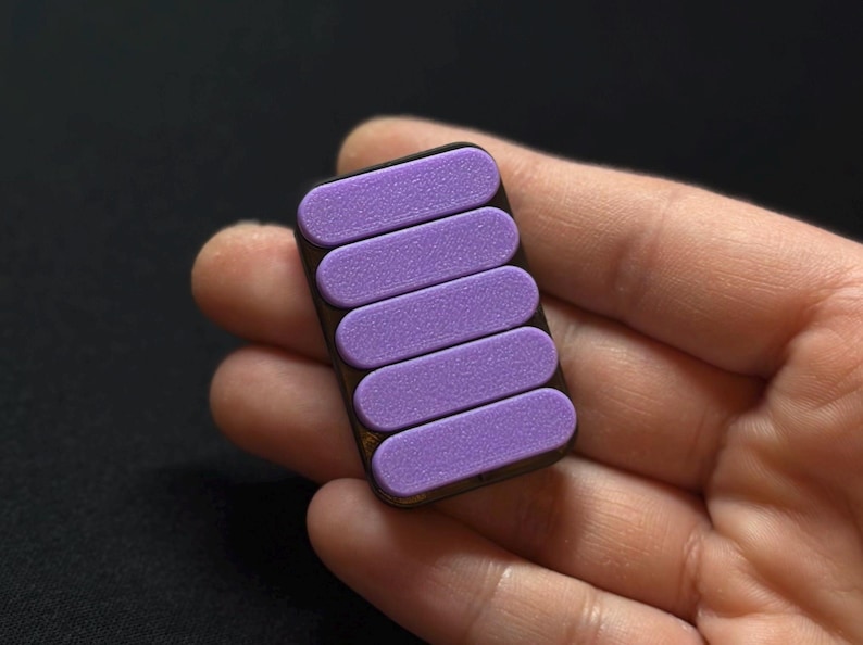 May include: A rectangular, dark brown object with five parallel, light purple, textured, rectangular pads. The object is held in a person's hand against a dark background. The pads are evenly spaced.