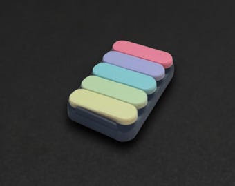 May include: A rectangular plastic case containing five stacked, pill-shaped adhesive markers in pastel colors. The markers are arranged in a vertical row, featuring shades of pink, purple, blue, green, and yellow, all housed within a dark gray base.