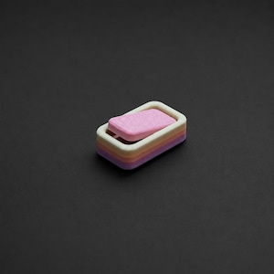 Può includere: Un portasapone multistrato con una saponetta rosa. Il portasapone ha strati di bianco, giallo e viola. La saponetta è rettangolare e rosa. Lo sfondo è nero pieno.