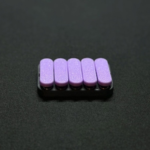 May include: A set of five lavender-colored nail files in a black holder. The files are rectangular with rounded edges and a textured surface. The holder is a small, rectangular tray.