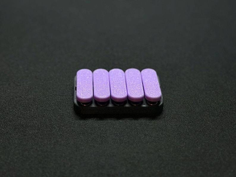 May include: A set of five lavender-colored nail files in a black holder. The files are rectangular with rounded edges and a textured surface. The holder is a small, rectangular tray.