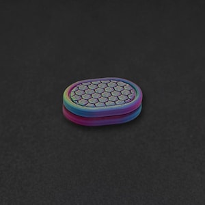 Op de afbeelding: Een ovaalvormig fidget-speeltje met een levendige, iriserende afwerking die verschuift tussen tinten blauw, paars en groen. Het bovenoppervlak toont een verhoogd, herhalend zeshoekig honingraatpatroon met een gladde, afgeronde rand.