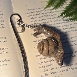 Könnte beinhalten: Dekorativer Metall-Lesezeichen mit einem Schneckenanhänger. Das Lesezeichen hat eine silberne Oberfläche mit einem detaillierten Design, und der Anhänger zeigt eine detaillierte Schneckenfigur mit Mond und Sternen.
