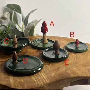 Puede incluir: Una colección de portainciensos de cerámica con forma de seta. Los soportes tienen bases verdes y negras. Algunas setas tienen sombreros rojos con lunares blancos, otras son marrones. Presentados sobre una superficie de madera.
