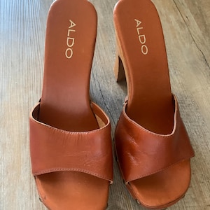 Könnte beinhalten: Ein Paar braune ALDO Clogs aus Leder mit Holzabsätzen. Die Sandalen mit offener Spitze haben einen breiten Riemen über dem Fuß, und der Markenname ist auf der Innensohle aufgedruckt.