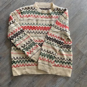 May include: A cream-coloured long-sleeve jumper featuring a repeating Fair Isle pattern in red, dark green, and dark brown. The knit garment has a crew neckline, ribbed cuffs, and a ribbed hem, with the patterned design across the chest and sleeves.