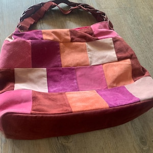May include: A patchwork tote bag with a brown leather strap. The bag is made of different shades of pink, red, and orange suede.