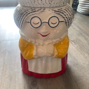 Puede incluir: Un tarro de galletas de cerámica con forma de anciana sonriente con gafas. El tarro presenta un gorro blanco, pelo gris y un vestido rojo. Los brazos de la mujer están cruzados y tiene un chal amarillo.