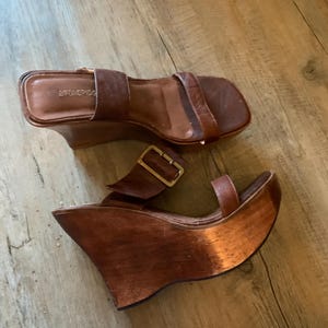 Pode incluir: Um par de sandálias de cunha de couro castanho com plataformas de madeira. As sandálias apresentam um design de bico quadrado, fecho de fivela e uma tira na parte superior do pé. O salto de cunha de madeira oferece suporte.