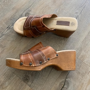 Op de afbeelding: Een paar bruine leren instapsandalen met een houten sleehak. Het bovenwerk heeft bruine leren bandjes in verschillende tinten met witte contraststiksels en kleine metalen klinknagels. Het beige voetbed is bij de hiel bedrukt met het cijfer 8.