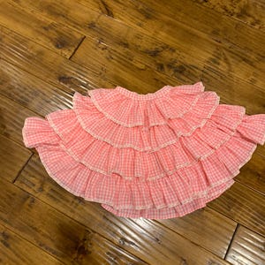 Puede incluir: Falda rosa y blanca de cuadros vichy con múltiples capas con volantes. La falda tiene un ribete de encaje blanco a lo largo de los bordes de cada volante. La tela parece ser de algodón ligero, adecuada para climas cálidos.