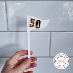 May include: White acrylic cake topper with a gold 50 flag. The topper is held in a hand. The background is a white tiled wall. The logo "Roo & Lou Design" is visible in the bottom right corner.