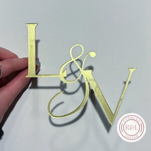 May include: Gold-coloured metal cake topper with the letters L & V and an ampersand in a cursive font. The topper is on a white background. The text "R&L DESIGN" is visible in a circle at the bottom of the image.
