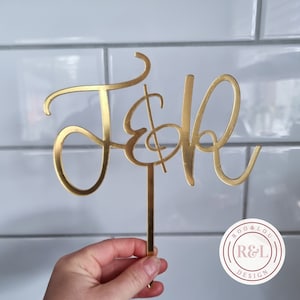 May include: Gold metal cake topper featuring the letters "J & B" in a cursive script font. The topper is mounted on a stick and is being held by a hand. The image also includes a logo for "Roo & Lou Design" in the bottom right corner.