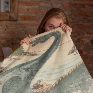 May include: A woman is partially hidden behind a cream-colored blanket with a blue and green illustration of a sea serpent. The blanket has a rope border and a Japanese-style design.