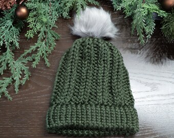 May include: A dark green knitted beanie with a fluffy white pom pom on top. The beanie is made of a thick, textured yarn and has a ribbed cuff.