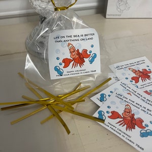 Può includere: Un sacchetto di plastica trasparente riempito con legacci dorati e decorato con piccole carte quadrate bianche con un'immagine di Sebastian de La Sirenetta. Le carte hanno il testo "La vita in mare è meglio di qualsiasi cosa sulla terra" e "Buon viaggio! Dai tuoi amici della cabina 7548".