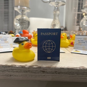 Può includere: Un'anatra di gomma gialla con un cappello da pirata nero e un passaporto blu con un globo sul coperchio. Il passaporto dice "PASSPORT" in lettere dorate.