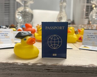 Puede incluir: Un pato de goma amarillo con un sombrero de pirata negro y un pasaporte azul con un globo terráqueo en la portada. El pasaporte dice "PASSPORT" en letras doradas.