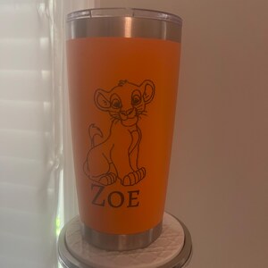 May include: Orange tumbler featuring a black line drawing of a lion cub and the name "ZOE". The tumbler has a stainless steel rim and lid. The tumbler is sitting on a decorative stand.