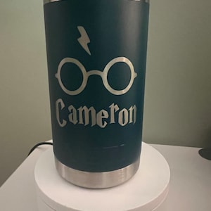 May include: A dark green travel mug with a silver lid and base. The mug has a silver lightning bolt and glasses design with the name "Cameron" engraved on it.