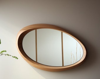 May include: A wooden oval-shaped mirror with a light wood frame. The mirror is hanging on a light beige wall.
