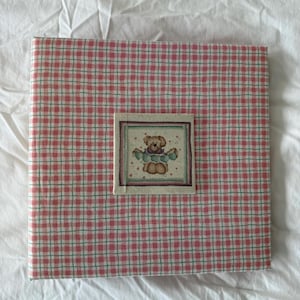 Può includere: Un album fotografico quadrato con un motivo a quadri rossi e bianchi. Una piccola immagine quadrata di un orsacchiotto è centrata sul davanti. L'album è su una superficie bianca.