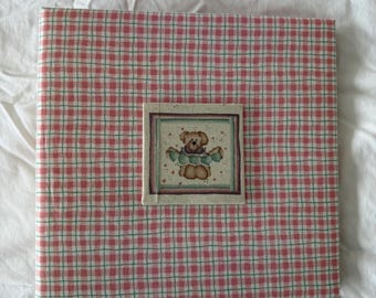 Puede incluir: Un álbum de fotos cuadrado con un patrón de cuadros rojos y blancos. Una pequeña imagen cuadrada de un oso de peluche está centrada en la parte delantera. El álbum está sobre una superficie blanca.