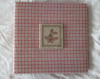 Puede incluir: Un álbum de fotos cuadrado con un patrón de cuadros rosa y blanco. Una pequeña imagen enmarcada de un conejito con un diseño floral está centrada en el álbum. El álbum está sobre una superficie blanca y texturizada.