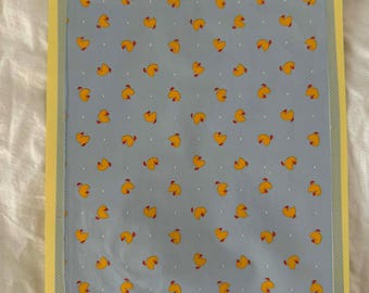 Puede incluir: Una impresión rectangular con un patrón de ilustraciones de patos amarillos sobre un fondo azul claro. La impresión está enmarcada con un borde amarillo y un patrón de puntos. Los patos tienen picos rojos y están espaciados uniformemente.