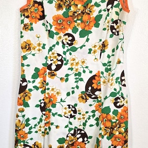 May include: Sleeveless white dress with a vibrant floral pattern in orange, yellow, and brown. The dress has a round neckline and orange trim. The dress is hanging on a blue hanger.