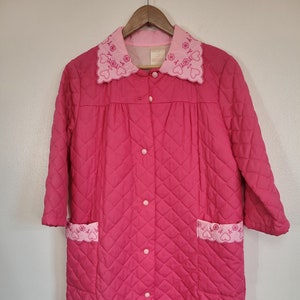 May include: A pink quilted robe with a white and pink embroidered collar and pockets. The robe has a button closure and long sleeves.