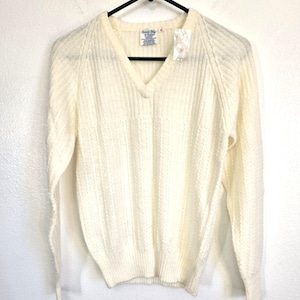 May include: Cream-colored V-neck sweater with long sleeves, hanging on a black hanger. The sweater has a ribbed knit texture and a tag attached near the neckline. The tag reads "Good Way by Fashion".