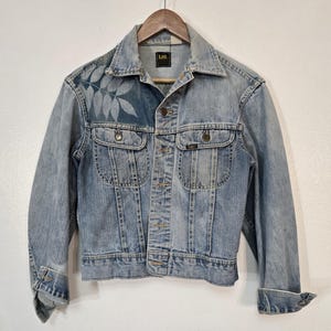 May include: A light blue denim jacket with a leaf design on the left side of the chest. The jacket has a button closure, two front pockets, and a Lee logo on the pocket flap.