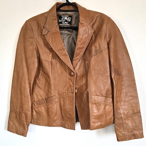 May include: A tan leather blazer with a notched collar, two front pockets, and a single-button closure. The jacket has a classic design and is hanging on a black hanger. The label reads "Genuine Leather, Made in Mexico."