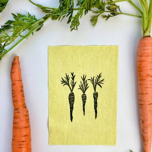 May include: An arrangement featuring fresh carrots and a small, light yellow fabric with a black print of three carrots. The carrots are orange with green tops, set against a white background.