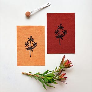 May include: Two rectangular paper sheets, one orange and one dark red, each featuring a black botanical print of a flowering plant. A silver measuring spoon containing a small amount of brown powder and a green plant sprig are also present.