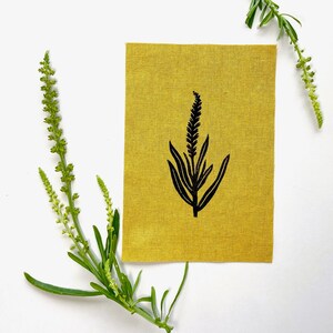 May include: A mustard yellow fabric square with a black botanical illustration of a plant. Green sprigs with tiny flowers are arranged around the fabric on a white background. The fabric has a textured appearance.