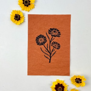 May include: A brown fabric patch with a black silhouette of three flowers with stems and leaves.
