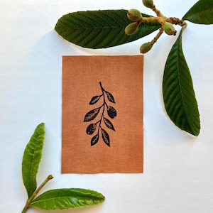 May include: A rectangular, rust-colored card with a black botanical illustration of a branch with leaves and berries. Green leaves and a branch with small buds are arranged around the card on a white background.