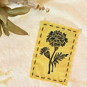 May include: A yellow fabric patch with a black floral design, featuring a large flower and smaller buds. The patch is outlined with black stitching. Dried leaves and flowers are in the background.