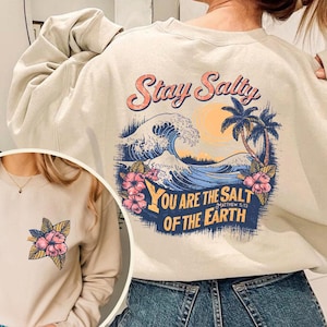 May include: A beige sweatshirt with a graphic print of a wave, palm trees, and the text "Stay Salty" and "You are the salt of the earth" on the back. The front of the sweatshirt has a small floral print.
