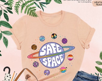 Op de afbeelding: Een heideperzikkleurig t-shirt met een afbeelding van een planeet met een ring eromheen. De planeet heeft de woorden "SAFE SPACE" erop geschreven in regenboogkleuren. De afbeelding is omringd door kleinere planeten met verschillende kleuren en patronen.