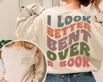 May include: A beige sweatshirt with the text "I LOOK BETTER BENT OVER A BOOK" in colorful, retro-style lettering. The sweatshirt is shown on a model wearing blue jeans.