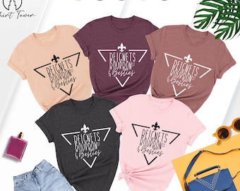 May include: Five t-shirts in different colors with a graphic design of a fleur-de-lis in a triangle with the text "Beignets Bourbon & Besties".